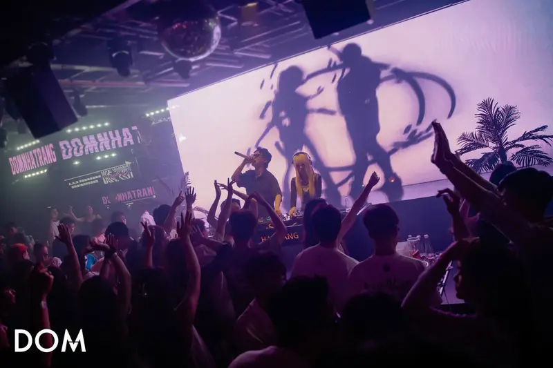 DJ và MC trước màn LED tím chiếu bóng người đang nhảy, bảng chữ DOM NHA TRANG treo trên giàn thép