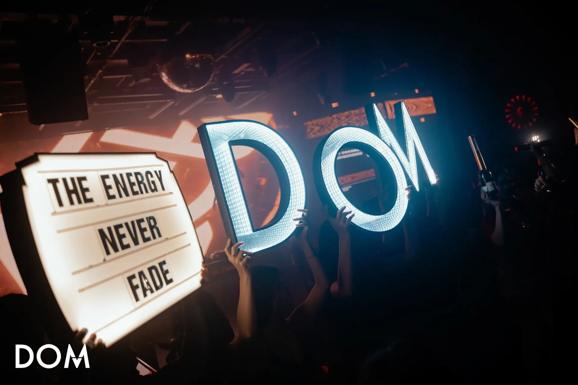 Khách giơ cao chữ DOM phát sáng và bảng đèn The Energy Never Fade giữa sàn